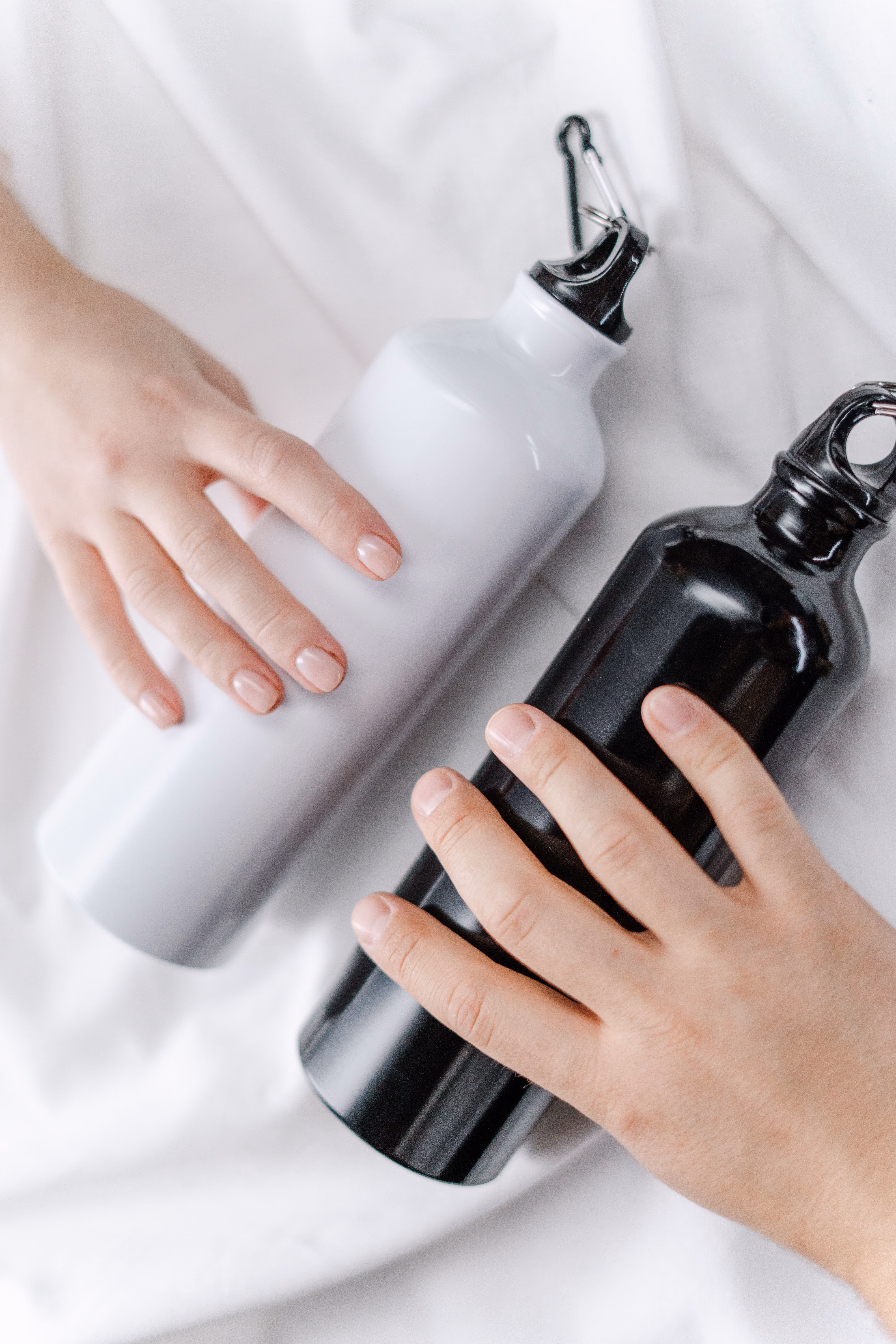 Two hands holding two aluminum water bottles, one white and one black, placed on a white fabric surface.