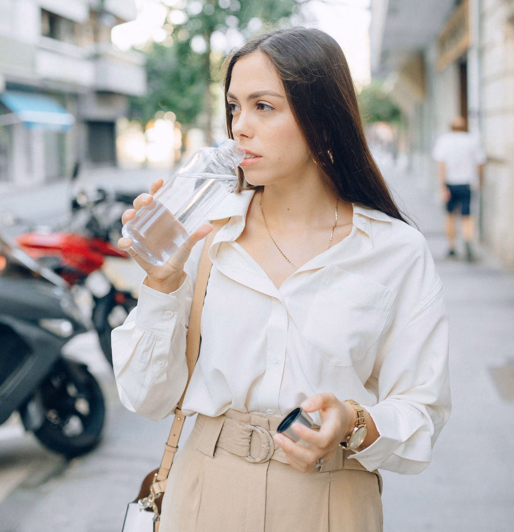 Young woman with long dark hair drinking water from a clear bottle on a city sidewalk, wearing a white blouse and beige high-waisted pants with a belt.